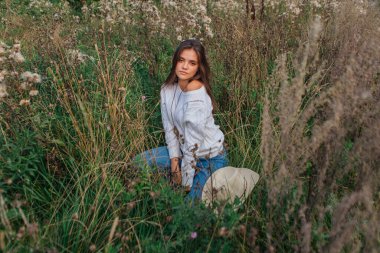 Güzel esmer genç kız beyaz kazak, kot pantolon ve kovboy hasır şapkası içinde kuru kahverengi çimlerin üzerinde oturuyor, gün batımında gülümsüyor ve gülüyor.