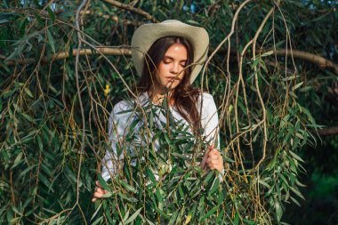 Beyaz kazak, kot pantolon ve kovboy hasır şapkası giymiş, yeşil yapraklı ağaç brançlarının arkasında duran güzel esmer kız.