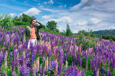 Yeşil ceketli uzun boylu yakışıklı bir adam lupin çiçeği tarlasında duruyor, doğanın güzelliğini emrediyor. Mor ve pembe lupinlerle çevrili bir adam..