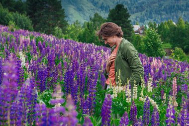 Yeşil ceketli uzun boylu yakışıklı bir adam lupin çiçeği tarlasında duruyor, doğanın güzelliğini emrediyor. Mor ve pembe lupinlerle çevrili bir adam..