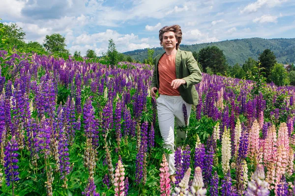 Tall handsome man in a green jacket standing on lupine flowers field, enjoing the beauty of nature. Man surrounded by purple and pink lupines.