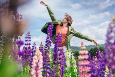 Yeşil ceketli uzun boylu yakışıklı bir adam lupin çiçeği tarlasında duruyor, doğanın güzelliğini emrediyor. Mor ve pembe lupinlerle çevrili bir adam..