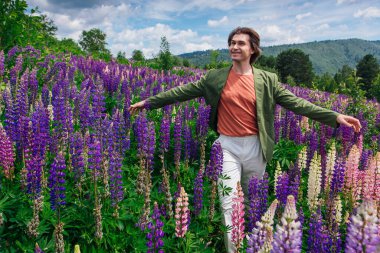 Yeşil ceketli uzun boylu yakışıklı bir adam lupin çiçeği tarlasında duruyor, doğanın güzelliğini emrediyor. Mor ve pembe lupinlerle çevrili bir adam..