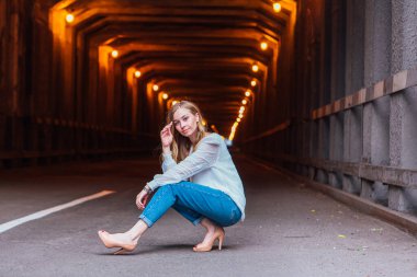Uzun sarı saçlı genç kız karanlık endüstriyel tünelin yanında oturuyor.