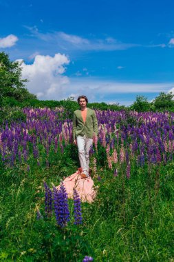 Yeşil ceketli uzun boylu yakışıklı bir adam lupine çiçekleri tarlasında pembe bir bez üzerinde yürüyor ve doğanın güzelliğini emrediyor. Mor ve pembe lupinlerle çevrili bir adam..