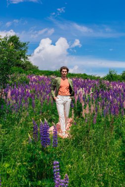 Yeşil ceketli uzun boylu yakışıklı bir adam lupine çiçekleri tarlasında pembe bir bez üzerinde yürüyor ve doğanın güzelliğini emrediyor. Mor ve pembe lupinlerle çevrili bir adam..