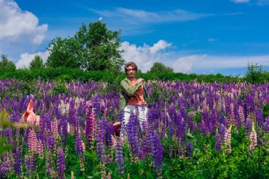 Yeşil ceketli uzun boylu yakışıklı bir adam lupin çiçeği tarlasında duruyor pembe bez atıyor ve doğanın güzelliğini buyuruyor. Mor ve pembe lupinlerle çevrili bir adam..