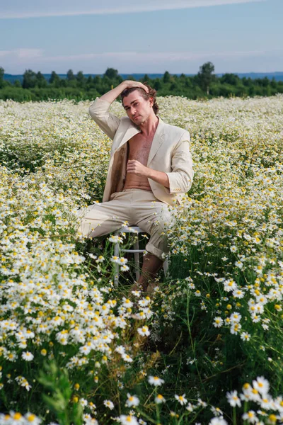 背の高いハンサムな男の服を着た白いスーツで裸の体に座っている上に白い椅子でカモミールの花のフィールド — ストック写真