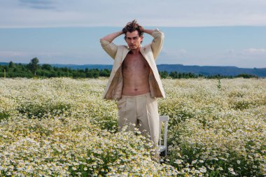 Beyaz takım elbiseli uzun boylu, yakışıklı bir adam. Üzerinde çıplak vücut, papatya çiçeği tarlasında duruyor.