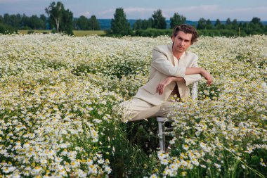 Beyaz takım elbiseli uzun boylu, yakışıklı bir adam papatya çiçeği tarlasında beyaz bir sandalyede oturuyor.