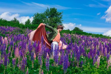 Yeşil ceketli uzun boylu yakışıklı bir adam lupin çiçekleri tarlasında duruyor pembe bir bezle, doğanın güzelliğini emrediyor. Mor ve pembe lupinlerle çevrili bir adam..