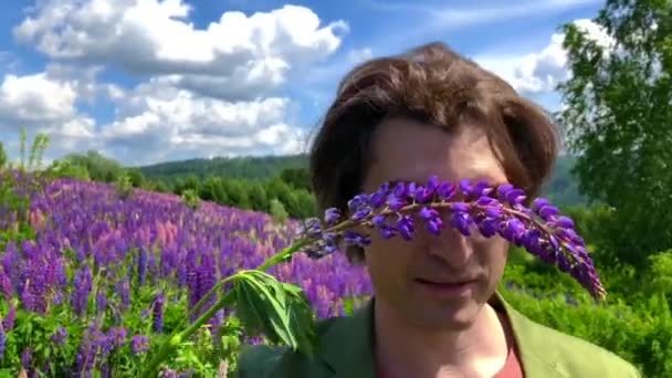 Високий красивий чоловік стоїть на полі квітів люпину з очима, закритими квіткою люпину — стокове відео
