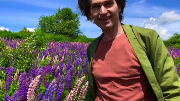 Alto bonito homem de pé no campo de flores de tremoço olhando para a câmera e sorrindo — Vídeo de Stock