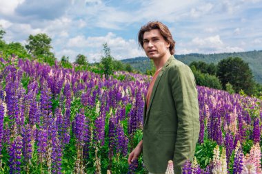 Yeşil ceketli uzun boylu yakışıklı bir adam lupin çiçeği tarlasında duruyor, doğanın güzelliğini emrediyor. Mor ve pembe lupinlerle çevrili bir adam..