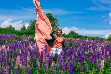 Yeşil ceketli uzun boylu yakışıklı bir adam lupin çiçeği tarlasında duruyor pembe bez atıyor ve doğanın güzelliğini buyuruyor. Mor ve pembe lupinlerle çevrili bir adam..