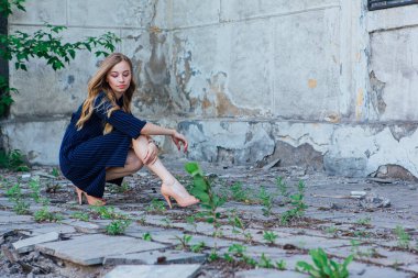 Gri duvarları olan eski bir binanın yanında oturan güzel sarışın bir kızın portresi.