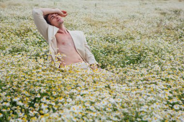 Beyaz takım elbiseli uzun boylu, yakışıklı bir adam papatya çiçeği tarlasında beyaz bir sandalyede oturuyor.