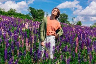 Yeşil ceketli uzun boylu yakışıklı bir adam lupin çiçeği tarlasında duruyor elinde lupin çiçekleri, doğanın güzelliğini emrediyor. Mor ve pembe lupinlerle çevrili bir adam..