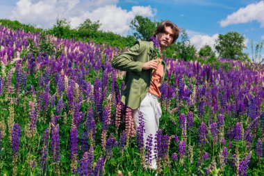 Yeşil ceketli uzun boylu yakışıklı bir adam lupin çiçeği tarlasında duruyor elinde lupin çiçekleri, doğanın güzelliğini emrediyor. Mor ve pembe lupinlerle çevrili bir adam..