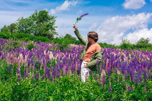 Yeşil ceketli uzun boylu yakışıklı bir adam lupin çiçeği tarlasında duruyor elinde lupin çiçekleri, doğanın güzelliğini emrediyor. Mor ve pembe lupinlerle çevrili bir adam..