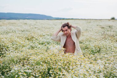 Beyaz takım elbiseli uzun boylu, yakışıklı bir adam papatya çiçeği tarlasında beyaz bir sandalyede oturuyor.