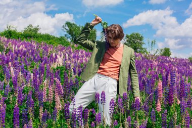 Yeşil ceketli uzun boylu yakışıklı bir adam lupin çiçeği tarlasında duruyor elinde lupin çiçekleri, doğanın güzelliğini emrediyor. Mor ve pembe lupinlerle çevrili bir adam..