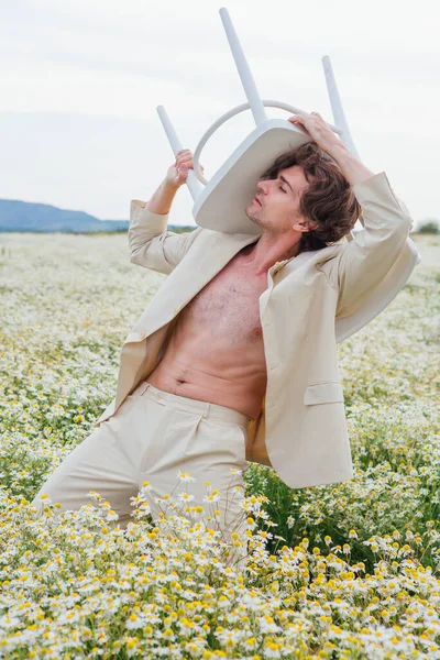 背の高いハンサムな男は白いスーツを着て カモミールの花のフィールドの真ん中に立って 彼の肩の上に白い椅子を保持 — ストック写真