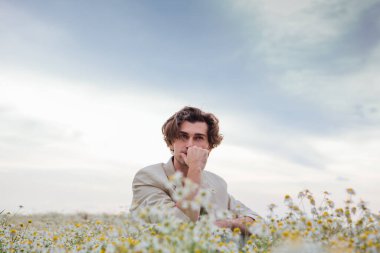 Beyaz takım elbiseli uzun boylu yakışıklı adam papatya çiçeği tarlasında beyaz bir sandalyede oturuyor.