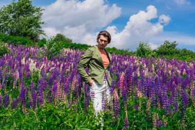 Yeşil ceketli uzun boylu yakışıklı bir adam lupin çiçeği tarlasında duruyor, doğanın güzelliğini emrediyor. Mor ve pembe lupinlerle çevrili bir adam..