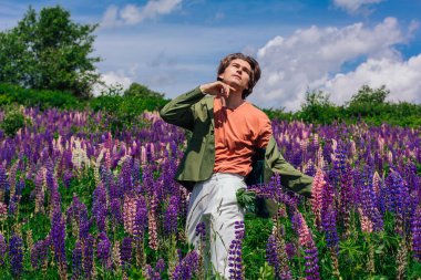 Yeşil ceketli uzun boylu yakışıklı bir adam lupin çiçeği tarlasında duruyor cebinde lupin çiçekleri, doğanın güzelliğini emrediyor. Mor ve pembe lupinlerle çevrili bir adam..