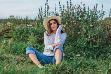 Beyaz kazak, kot pantolon ve kovboy hasır şapkası giyen genç, güzel esmer kadın yeşil çimlerin üzerinde oturuyor, gün batımında gülümsüyor ve gülüyor.