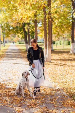 Altın sonbahar parkında Dalmaçyalı bir köpekle balerin. Beyaz balerin etekli ve siyah deri ceketli bir kadın. Sonbahar parkında benekli dalmaçyalı köpeğiyle dans ediyor..