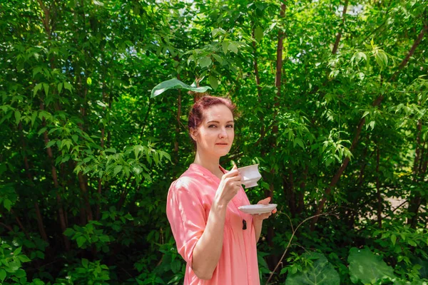 Red hair woman dressed in pink blouse with big green leaf in her hair drinking tea from the white cup in the blooming garden