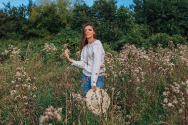 Beyaz kazak, kot pantolon ve kovboy hasır şapkası giymiş genç, güzel esmer kadın kuru kahverengi çimlerin üzerinde duruyor, gün batımında gülümsüyor ve gülüyor.