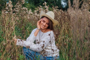 Beyaz kazak, kot pantolon ve kovboy hasır şapkası giyen genç, güzel esmer kadın kuru kahverengi çimlerin üzerinde oturuyor, gün batımında gülümsüyor ve gülüyor.