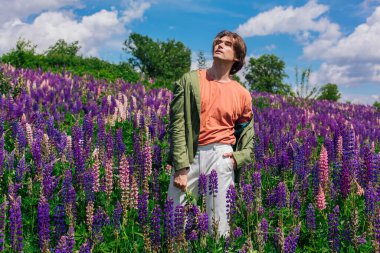 Yeşil ceketli uzun boylu yakışıklı bir adam lupin çiçeği tarlasında duruyor elinde lupin çiçekleri, doğanın güzelliğini emrediyor. Mor ve pembe lupinlerle çevrili bir adam..