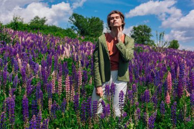 Yeşil ceketli uzun boylu yakışıklı bir adam lupin çiçeği tarlasında duruyor elinde lupin çiçekleri, doğanın güzelliğini emrediyor. Mor ve pembe lupinlerle çevrili bir adam..