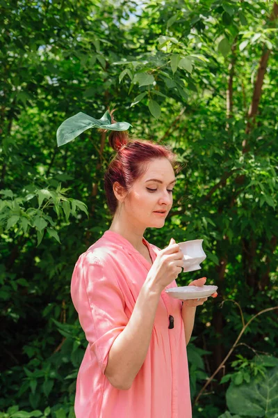 Red hair woman dressed in pink blouse with big green leaf in her hair drinking tea from the white cup in the blooming garden