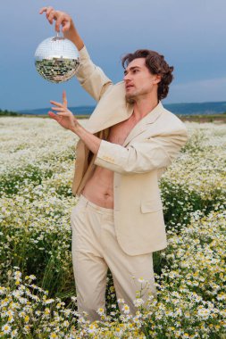 Beyaz takım elbiseli uzun boylu, çıplak bir adam papatya çiçeği tarlasında dikiliyor, elinde ayna disko topu tutuyor..