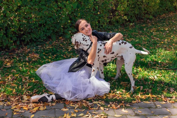 黄金の秋の公園でダルマチア犬とバレリーナ 女性バレリーナでホワイトバレエスカートと黒革のジャケットとでポイント靴で秋の公園抱擁彼女のきれいなダルマチア犬 — ストック写真