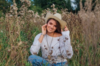 Beyaz kazak, kot pantolon ve kovboy hasır şapkası giyen genç, güzel esmer kadın kuru kahverengi çimlerin üzerinde oturuyor, gün batımında gülümsüyor ve gülüyor.