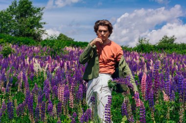 Yeşil ceketli uzun boylu yakışıklı bir adam lupin çiçeği tarlasında duruyor cebinde lupin çiçekleri, doğanın güzelliğini emrediyor. Mor ve pembe lupinlerle çevrili bir adam..