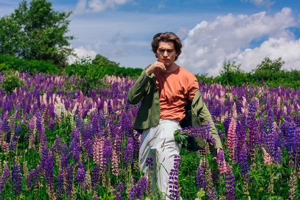 Tall handsome man in a green jacket standing on lupine flowers field with lupin flowers in his pocket, enjoing the beauty of nature. Man surrounded by purple and pink lupines.