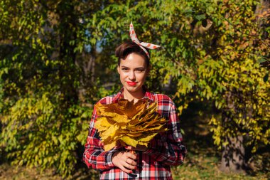 Makyajlı ve tokalı saçları olan güzel bir kadın güneşli bir sonbahar gününde büyük bir buket akçaağaç sarısı yaprağı tutuyor..