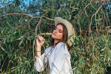 Beyaz kazak, kot pantolon ve kovboy hasır şapkası giymiş genç, güzel esmer kadın yeşil yapraklı ağaç brunch 'larına yakın duruyor.