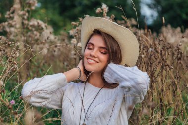 Beyaz kazak, kot pantolon ve kovboy hasır şapkası giyen genç, güzel esmer kadın kuru kahverengi çimlerin üzerinde oturuyor, gün batımında gülümsüyor ve gülüyor.