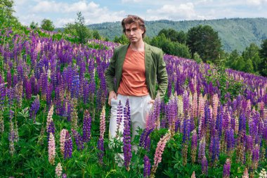 Yeşil ceketli uzun boylu yakışıklı bir adam lupin çiçeği tarlasında duruyor, doğanın güzelliğini emrediyor. Mor ve pembe lupinlerle çevrili bir adam..