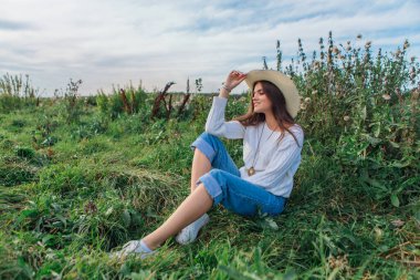 Beyaz kazak, kot pantolon ve kovboy hasır şapkası giyen genç, güzel esmer kadın yeşil çimlerin üzerinde oturuyor, gün batımında gülümsüyor ve gülüyor.