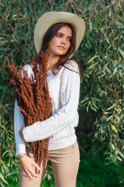 Beyaz kazak, kot pantolon ve kovboy hasır şapkası giymiş genç, güzel esmer kadın elinde kuru yapraklarla ağaç brunch 'larına yakın duruyor.