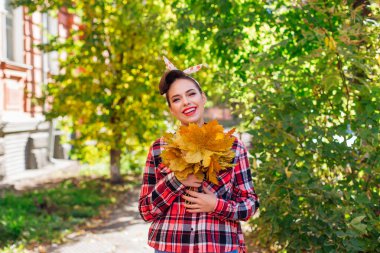 Makyajlı ve tokalı saçları olan güzel bir kadın güneşli bir sonbahar gününde büyük bir buket akçaağaç sarısı yaprağı tutuyor..